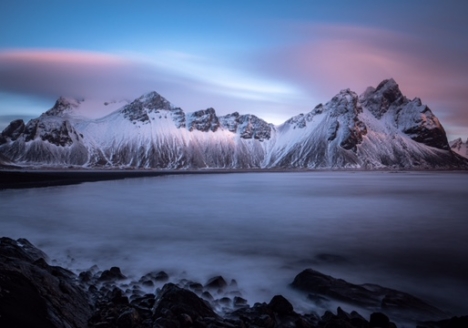 Schneebedeckte Berge und davor eine ruhige Wasserfläche aus der Nebel steigt Die Wolken über den Bergen sind von der Sonne leicht rosa angestellt. Symbol für Gustav Mahler